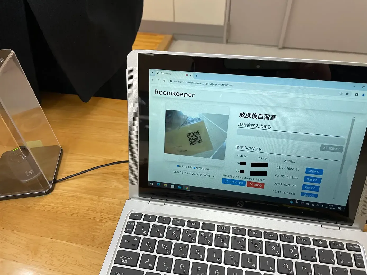 木製デスク上のノート PC 画面を写した写真。ブラウザには入退室管理システム「Roomkeeper」が表示され、画面左上に大見出し「Roomkeeper」、下にカメラ映像のプレビューエリアがあり、 QR コード付きカードを読み取り中であることがわかる。右側に「放課後自習室」という見出しがあり、「IDを直接入力する」入力欄と「滞在中のゲスト」の一覧を表示している表がある。下部には連続で同じゲストをスキャンするか確認するポップアップが表示されている。PC 左横には透明ケースに入った読み取りデバイスと USB ケーブルが見える。